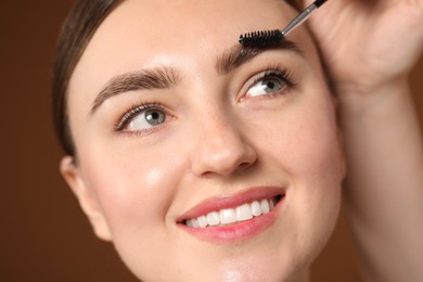 Photo of Cosmetologist brushing young woman's eyebrow with spoolie on brown background, closeup