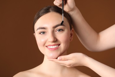 Photo of Cosmetologist brushing young woman's eyebrow with spoolie on brown background, closeup