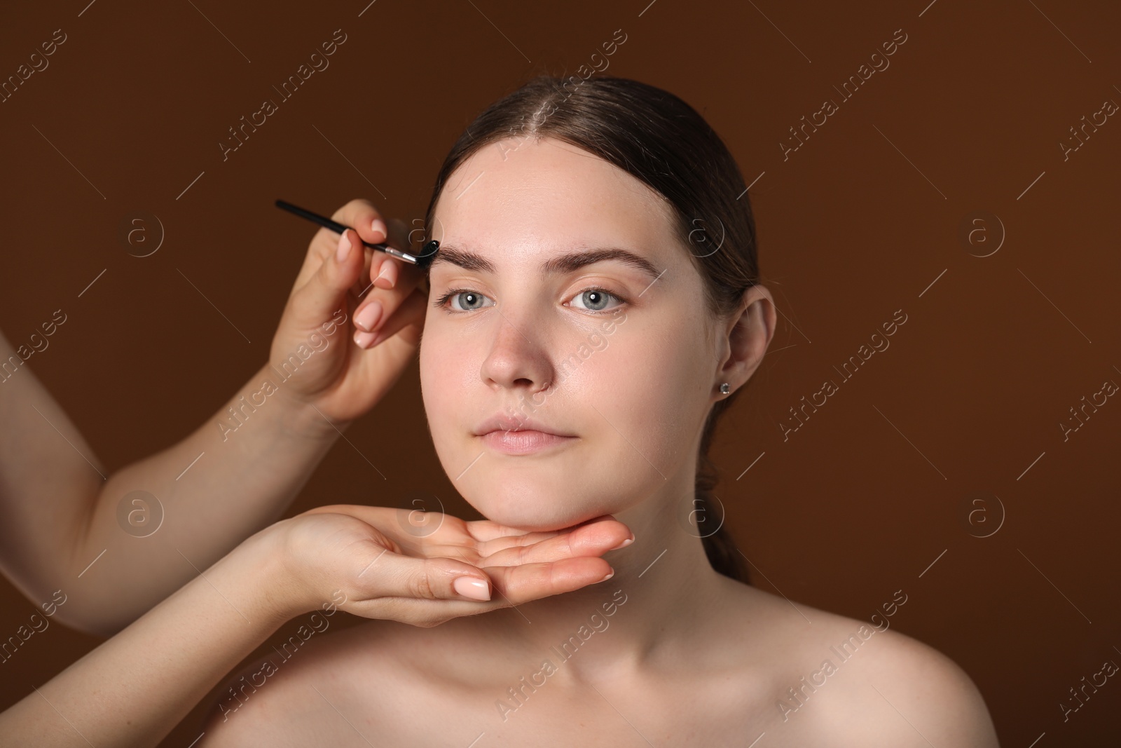 Cosmetologist brushing young woman's eyebrow with spoolie on brown background, closeup Photo of Cosmetologist brushing young woman's eyebrow with spoolie on brown background, closeup