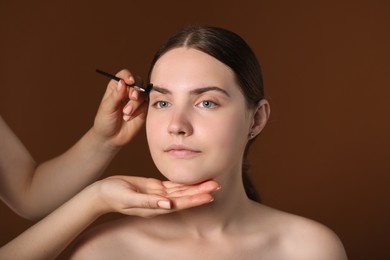 Photo of Cosmetologist brushing young woman's eyebrow with spoolie on brown background, closeup