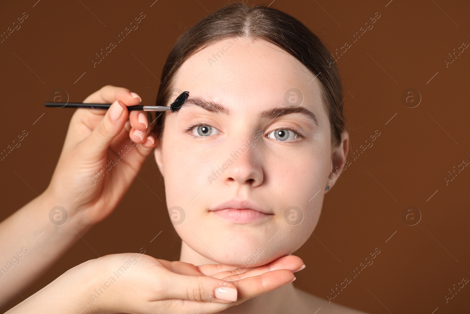 Cosmetologist brushing young woman's eyebrow with spoolie on brown background, closeup Photo of Cosmetologist brushing young woman's eyebrow with spoolie on brown background, closeup