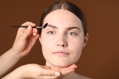 Photo of Cosmetologist brushing young woman's eyebrow with spoolie on brown background, closeup
