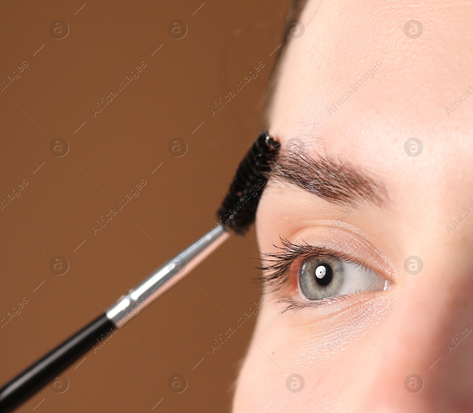 Photo of Young woman brushing her eyebrow with spoolie on brown background, closeup