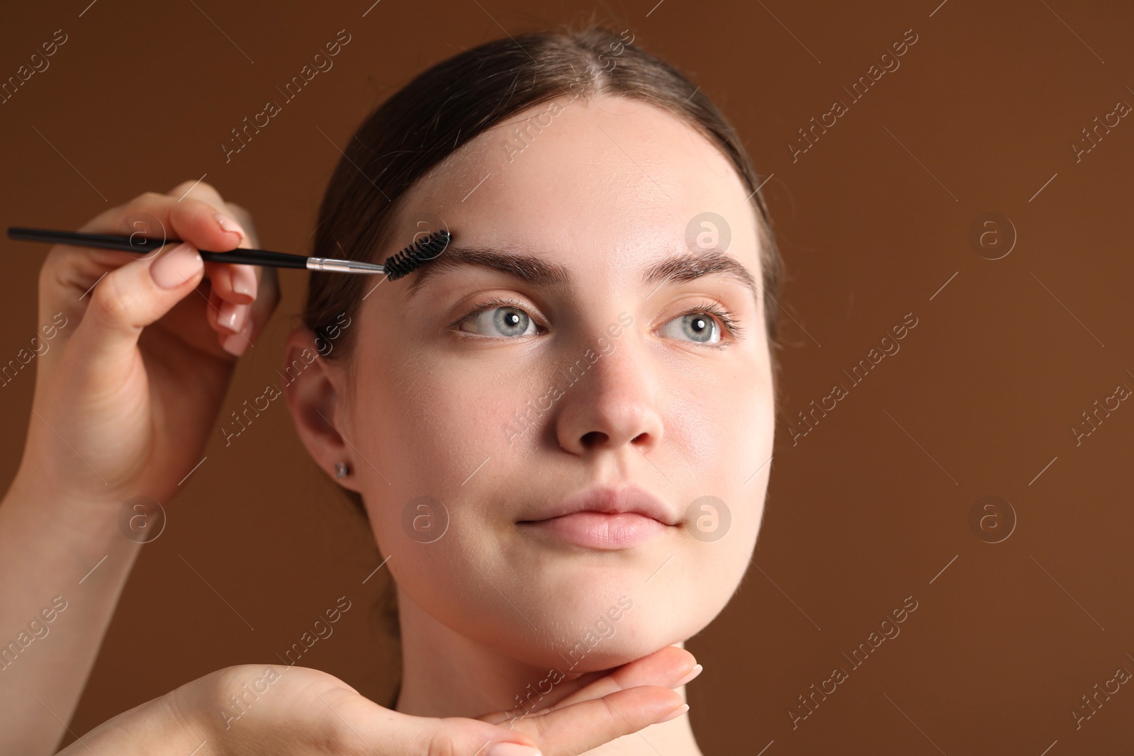 Cosmetologist brushing young woman's eyebrow with spoolie on brown background, closeup Photo of Cosmetologist brushing young woman's eyebrow with spoolie on brown background, closeup