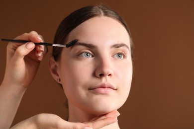 Photo of Cosmetologist brushing young woman's eyebrow with spoolie on brown background, closeup