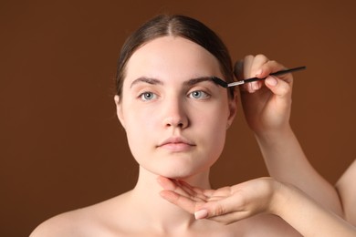 Photo of Cosmetologist brushing young woman's eyebrow with spoolie on brown background, closeup