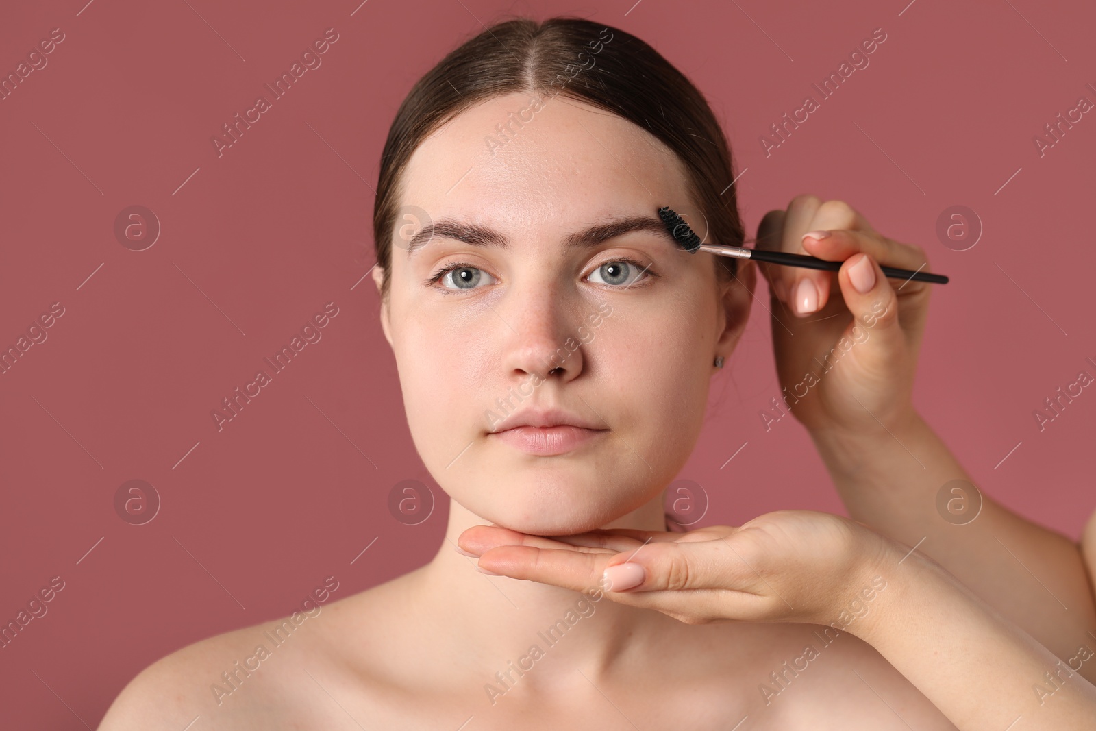 Cosmetologist brushing young woman's eyebrow with spoolie on pink background, closeup Photo of Cosmetologist brushing young woman's eyebrow with spoolie on pink background, closeup