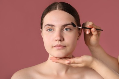 Photo of Cosmetologist brushing young woman's eyebrow with spoolie on pink background, closeup