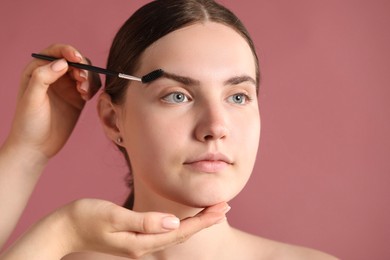 Photo of Cosmetologist brushing young woman's eyebrow with spoolie on pink background, closeup