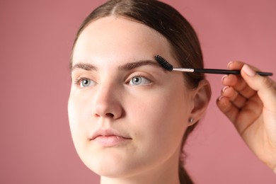 Photo of Cosmetologist brushing young woman's eyebrow with spoolie on pink background, closeup