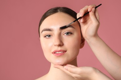 Photo of Cosmetologist brushing young woman's eyebrow with spoolie on pink background, closeup