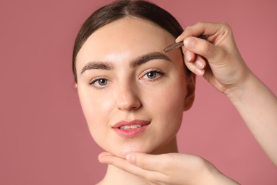 Photo of Cosmetologist plucking young woman's eyebrow with tweezers on pink background, closeup
