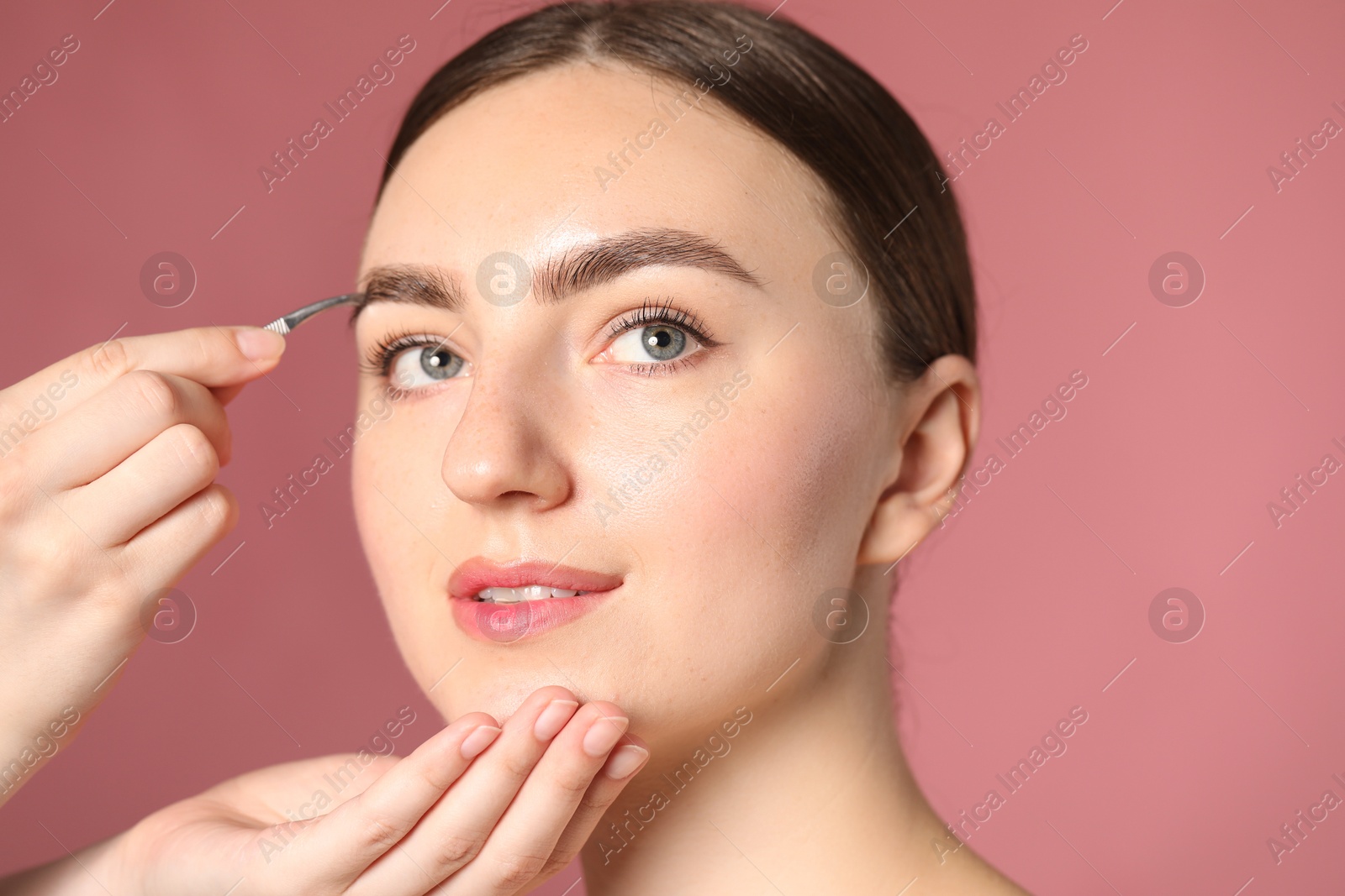 Cosmetologist plucking young woman's eyebrow with tweezers on pink background, closeup Photo of Cosmetologist plucking young woman's eyebrow with tweezers on pink background, closeup