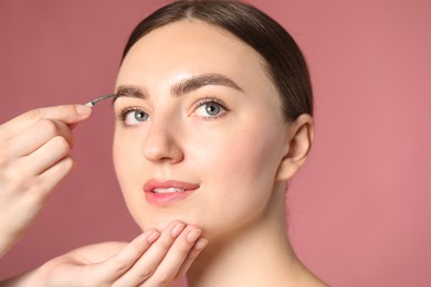 Photo of Cosmetologist plucking young woman's eyebrow with tweezers on pink background, closeup