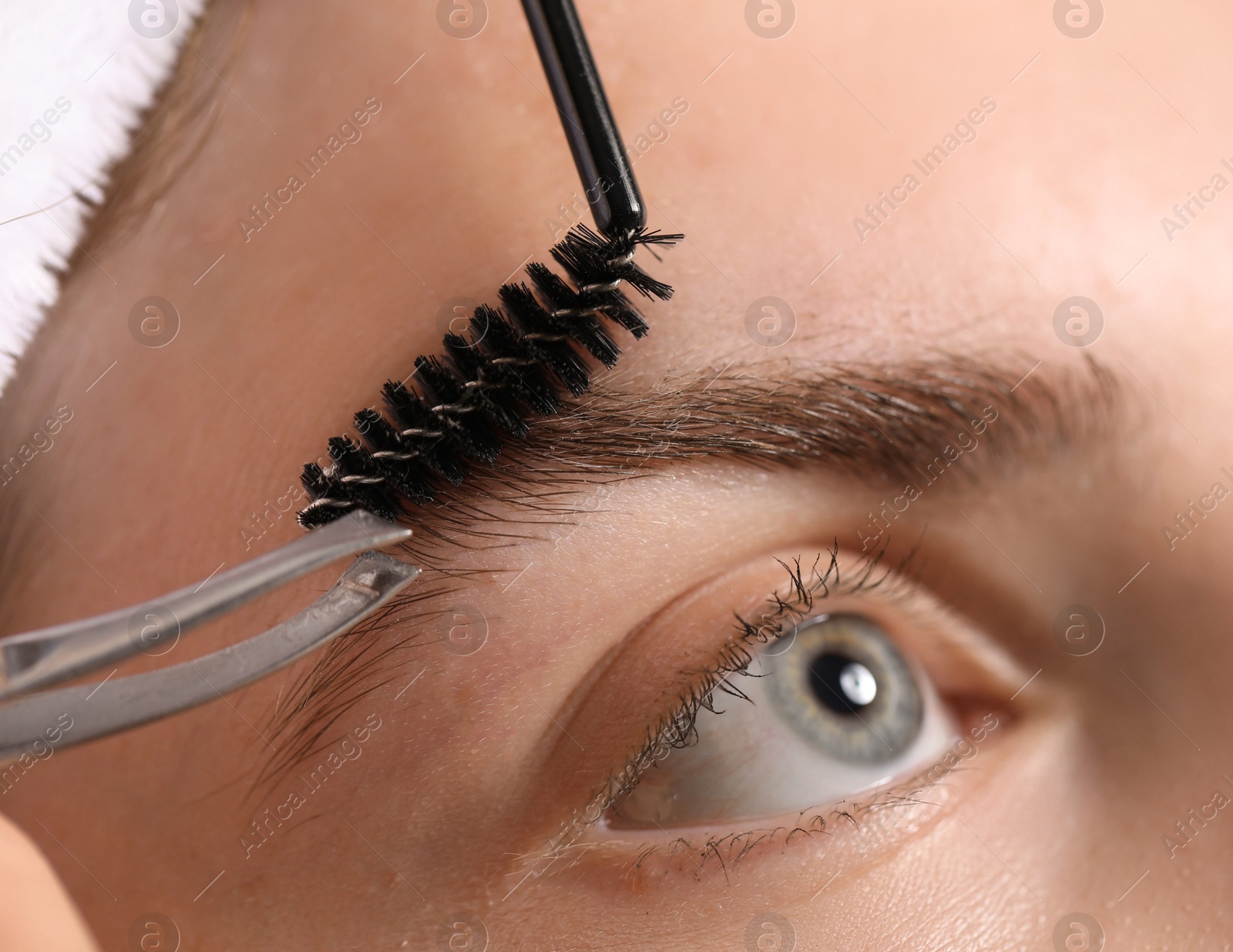 Photo of Cosmetologist plucking and brushing young woman's eyebrow, closeup