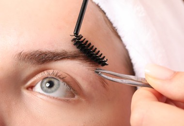Photo of Cosmetologist plucking and brushing young woman's eyebrow, closeup