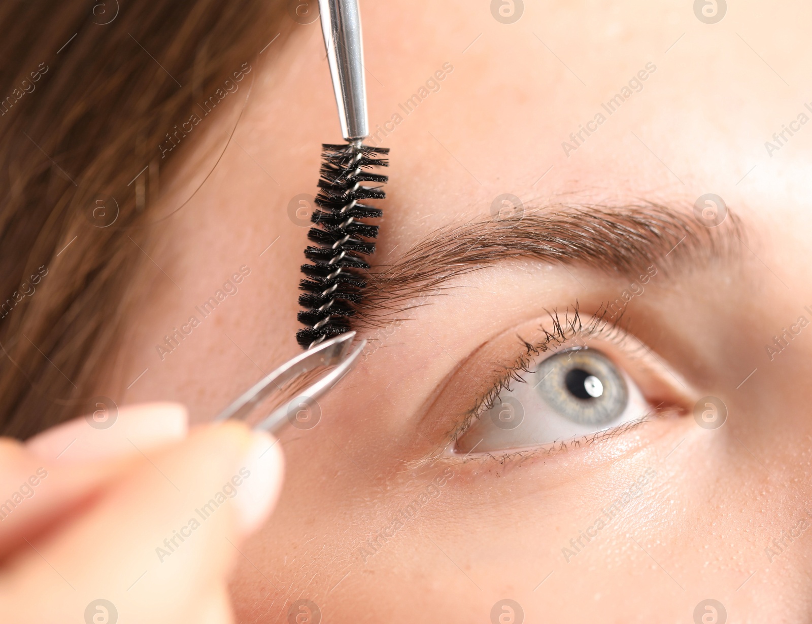 Photo of Cosmetologist plucking and brushing young woman's eyebrow, closeup
