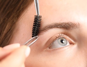Photo of Cosmetologist plucking and brushing young woman's eyebrow, closeup