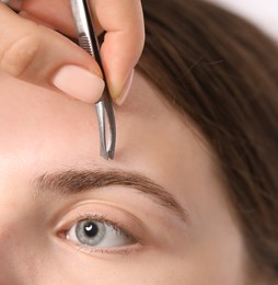 Photo of Cosmetologist plucking young woman's eyebrow with tweezers, closeup
