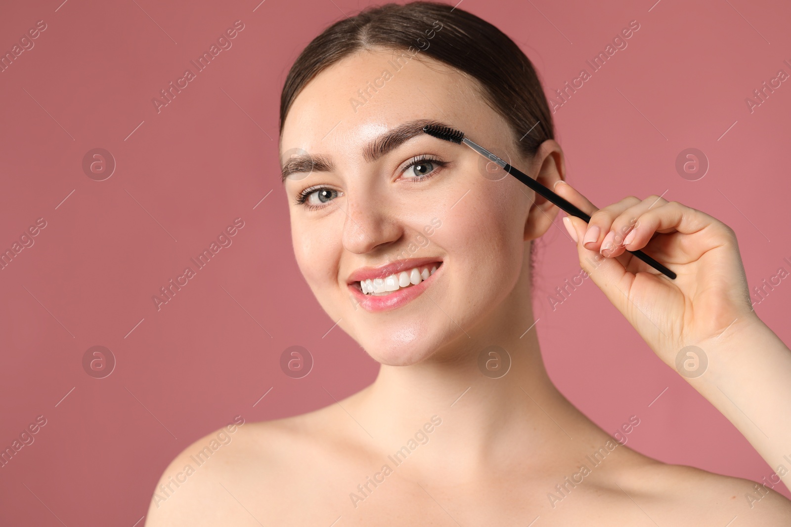 Young woman brushing her eyebrow with spoolie on pink background Photo of Young woman brushing her eyebrow with spoolie on pink background