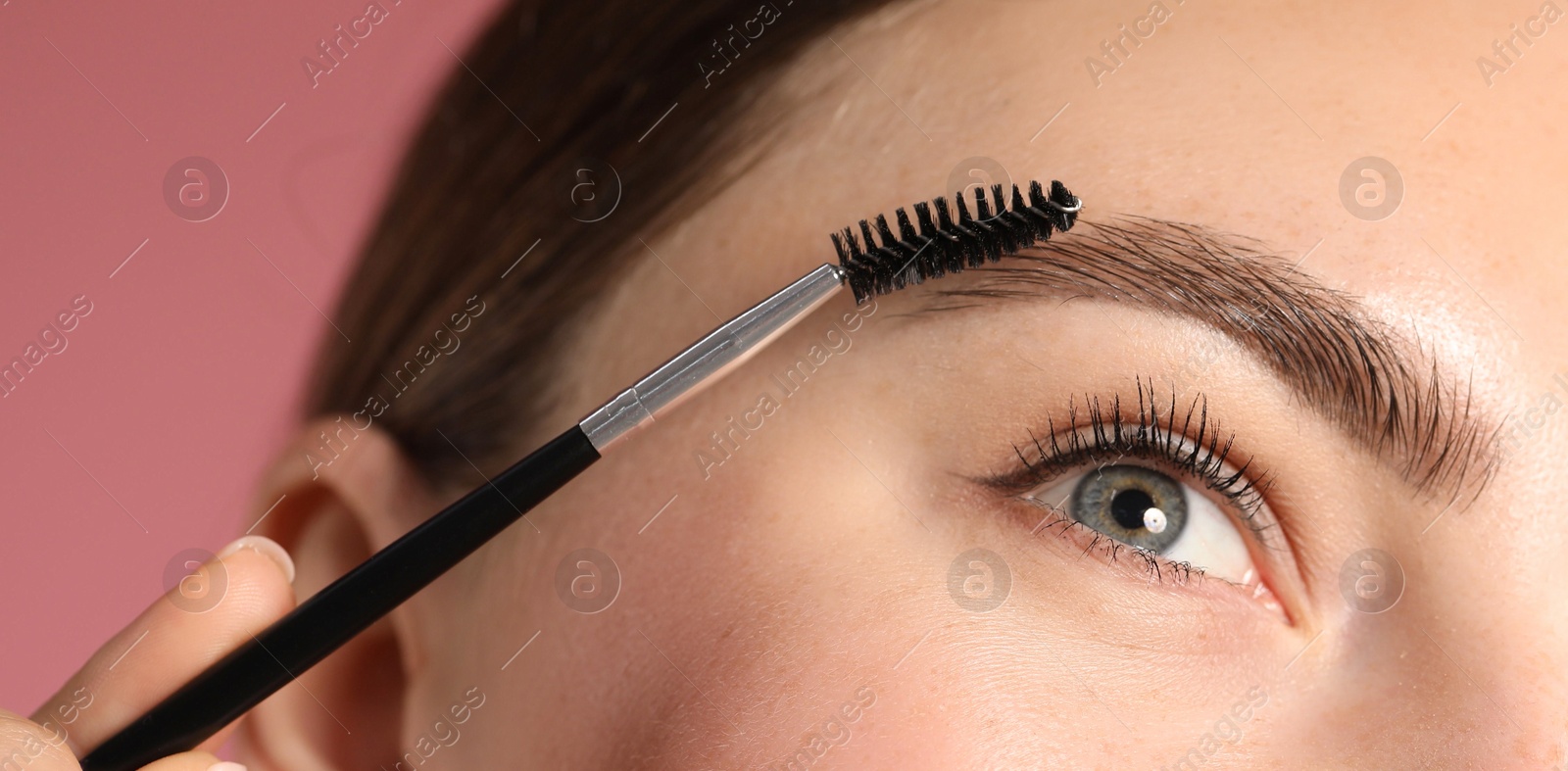 Photo of Young woman brushing her eyebrow with spoolie on pink background, closeup