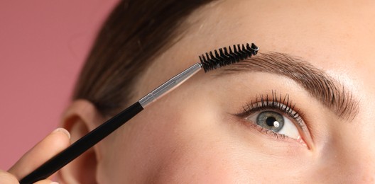 Photo of Young woman brushing her eyebrow with spoolie on pink background, closeup