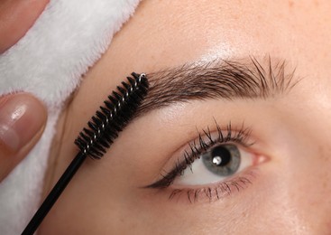 Photo of Cosmetologist brushing young woman's eyebrow with spoolie, closeup
