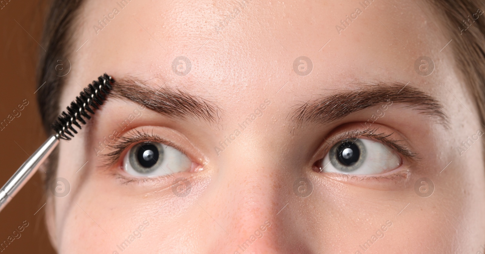 Photo of Young woman brushing her eyebrow with spoolie on brown background, closeup