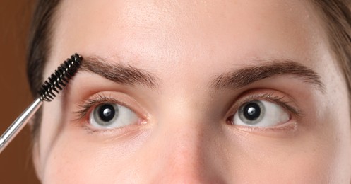 Photo of Young woman brushing her eyebrow with spoolie on brown background, closeup