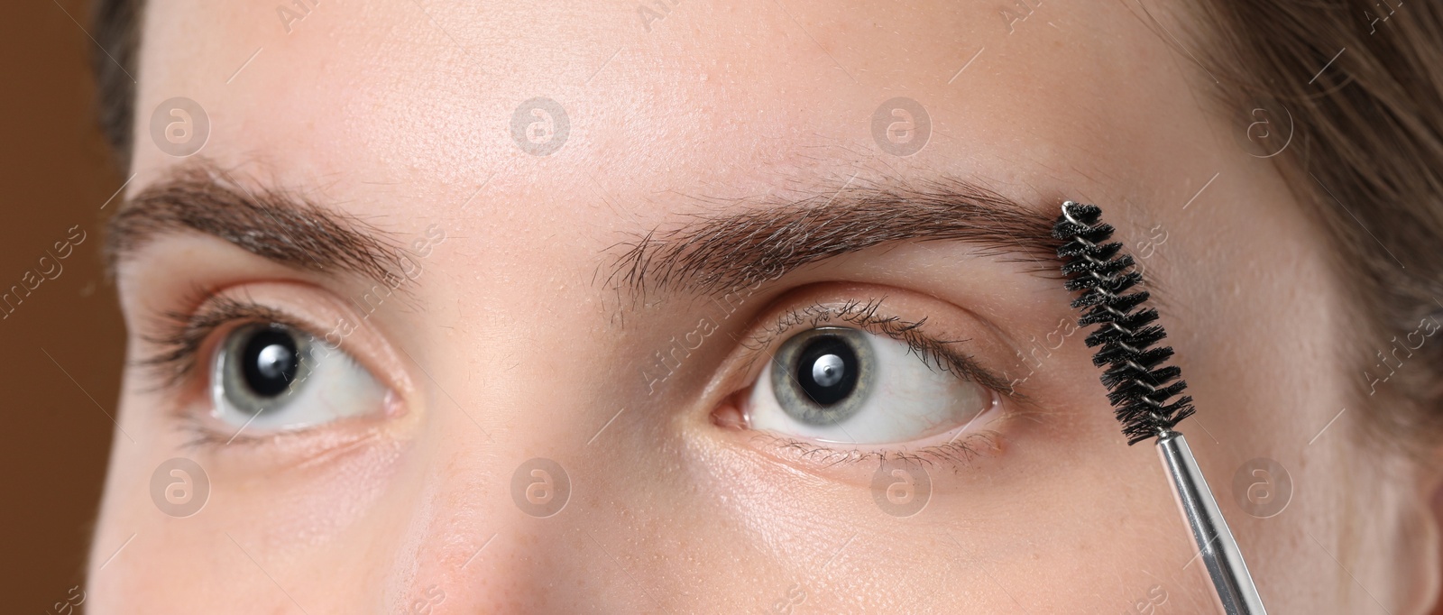 Photo of Young woman brushing her eyebrow with spoolie on brown background, closeup
