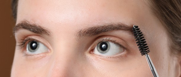 Photo of Young woman brushing her eyebrow with spoolie on brown background, closeup