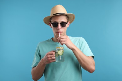 Photo of Man in sunglasses and hat drinking refreshing beverage from mason jar on light blue background