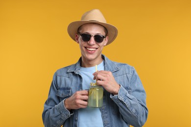 Photo of Happy man in hat and sunglasses with mason jar of lemonade on yellow background. Refreshing drink