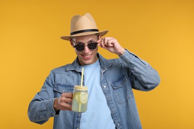 Photo of Happy man in hat and sunglasses with mason jar of lemonade on yellow background. Refreshing drink