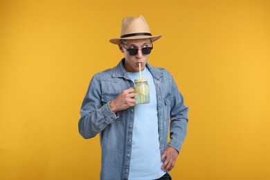 Photo of Man in sunglasses and hat drinking lemonade from mason jar on yellow background. Refreshing drink