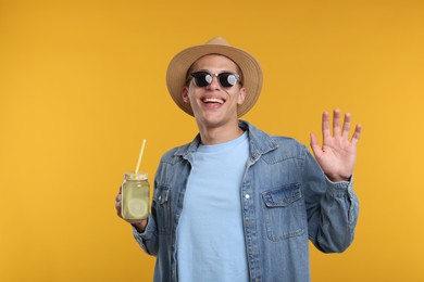 Photo of Happy man in hat and sunglasses with mason jar of lemonade on yellow background. Refreshing drink