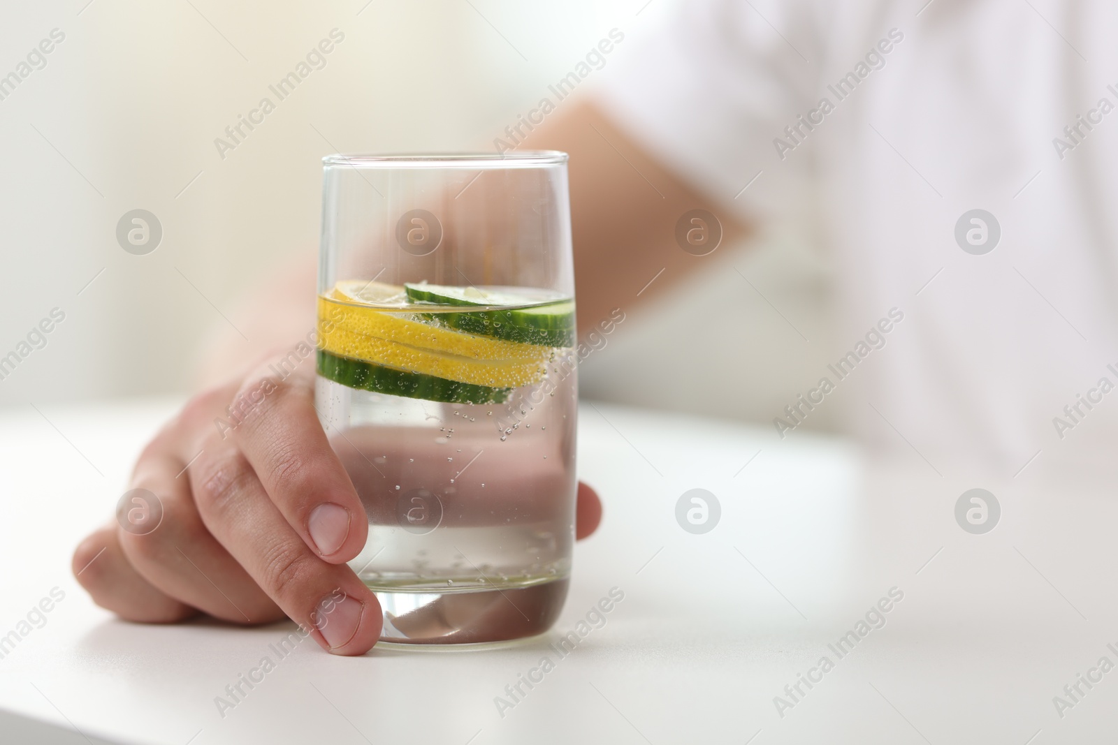 Photo of Man with glass of water with cucumber and lemon at white table indoors, closeup. Refreshing drink