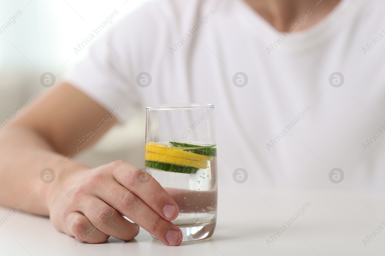 Photo of Man with glass of water with cucumber and lemon at white table indoors, closeup. Refreshing drink