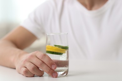 Photo of Man with glass of water with cucumber and lemon at white table indoors, closeup. Refreshing drink