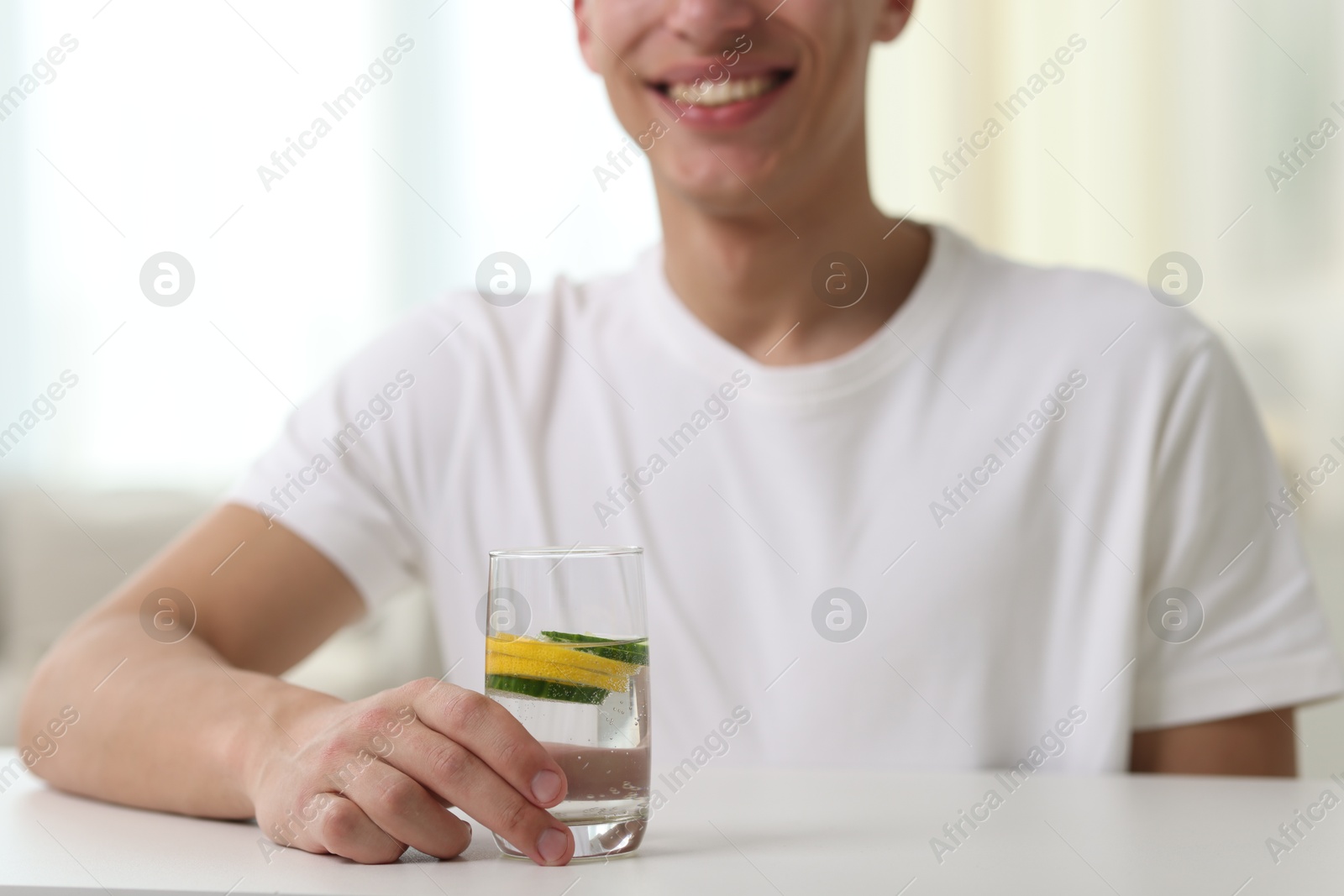 Smiling man with glass of water with cucumber and lemon at white table indoors, closeup. Refreshing drink Photo of Smiling man with glass of water with cucumber and lemon at white table indoors, closeup. Refreshing drink