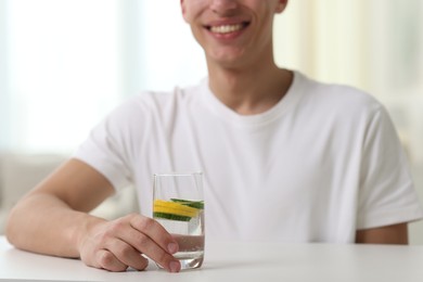 Photo of Smiling man with glass of water with cucumber and lemon at white table indoors, closeup. Refreshing drink