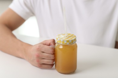 Photo of Man with mason jar of orange juice at white table indoors, closeup. Refreshing drink