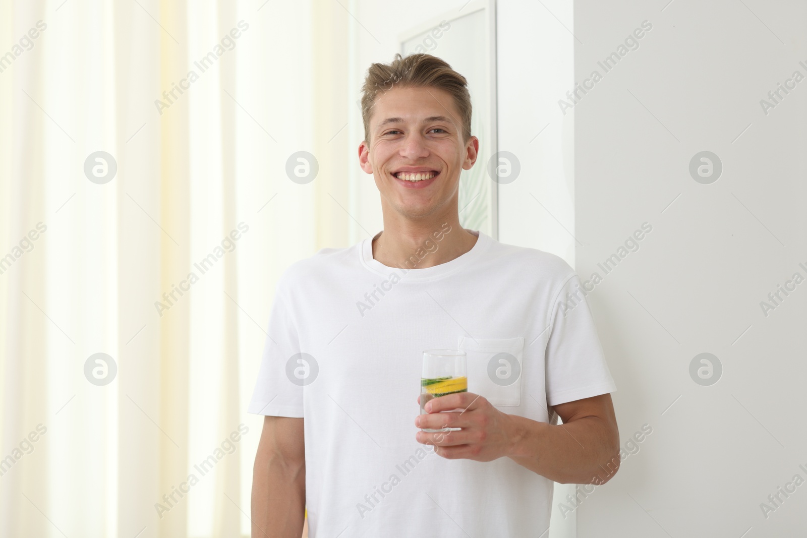 Smiling man with glass of water with cucumber and lemon near white wall indoors. Refreshing drink Photo of Smiling man with glass of water with cucumber and lemon near white wall indoors. Refreshing drink