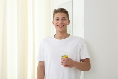 Photo of Smiling man with glass of water with cucumber and lemon near white wall indoors. Refreshing drink