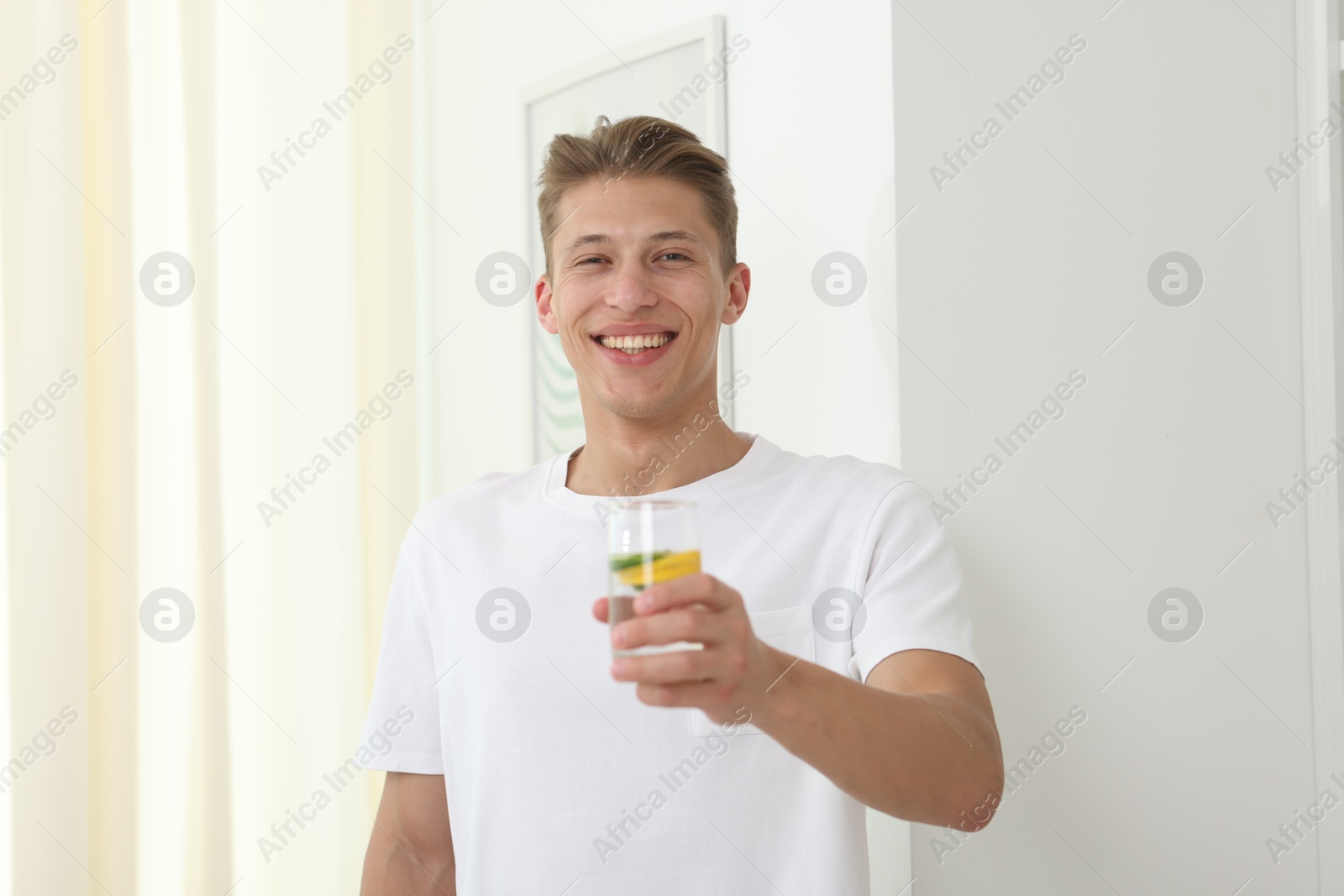 Smiling man with glass of water with cucumber and lemon near white wall indoors. Refreshing drink Photo of Smiling man with glass of water with cucumber and lemon near white wall indoors. Refreshing drink