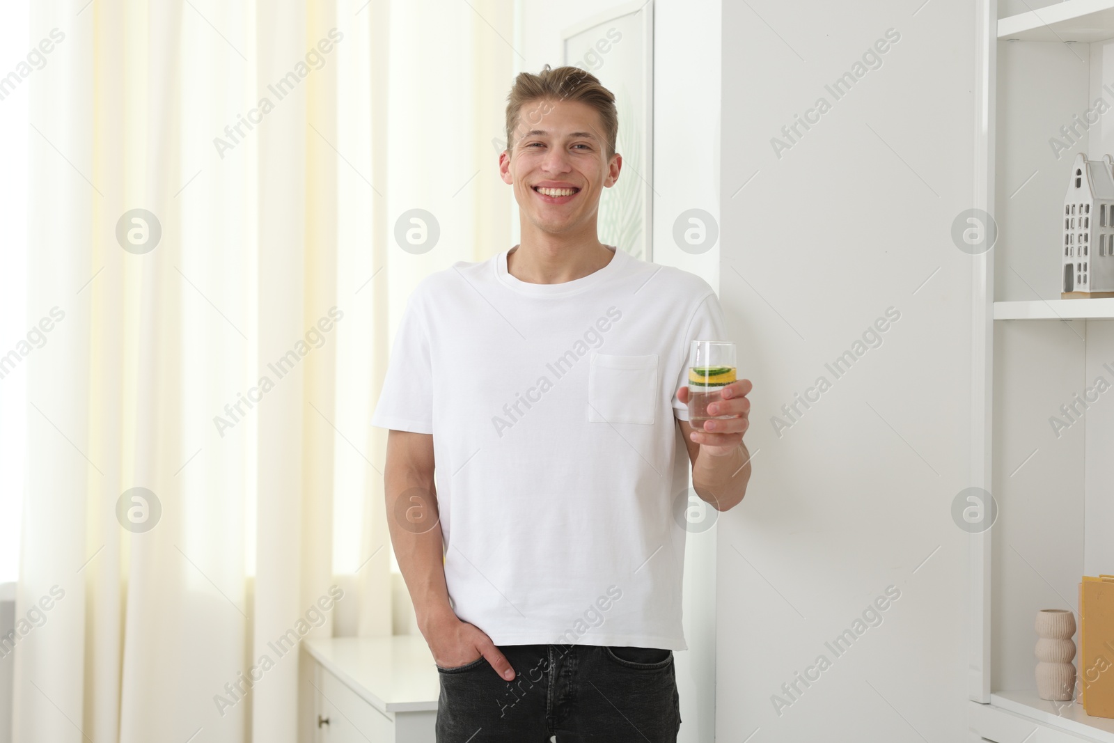 Smiling man with glass of water with cucumber and lemon near white wall indoors. Refreshing drink Photo of Smiling man with glass of water with cucumber and lemon near white wall indoors. Refreshing drink