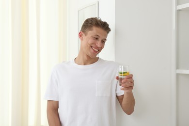 Photo of Smiling man with glass of water with cucumber and lemon near white wall indoors. Refreshing drink