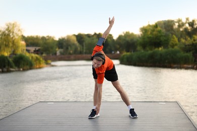 Photo of Young sportsman in headphones and armband doing morning exercise on pier near river