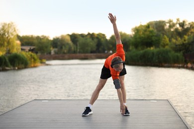 Photo of Young sportsman in headphones and armband doing morning exercise on pier near river