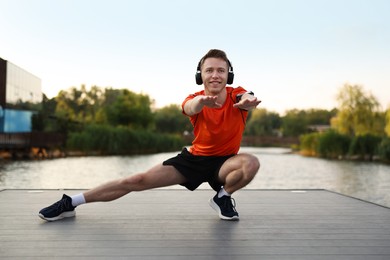 Photo of Smiling sportsman in headphones and armband doing morning exercise on pier near river